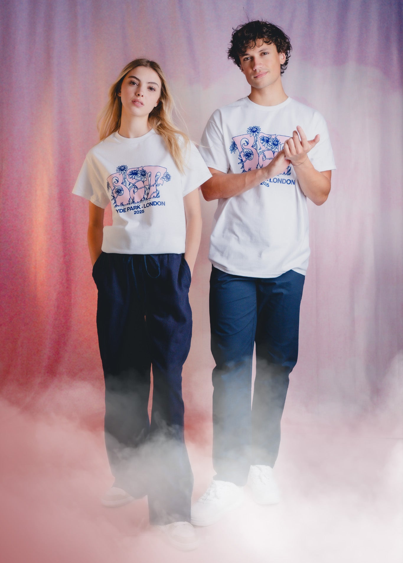 Two young adults stand in front of a pink and purple backdrop with mist at their feet, both wearing white graphic t-shirts and dark pants, gazing confidently at the camera.
