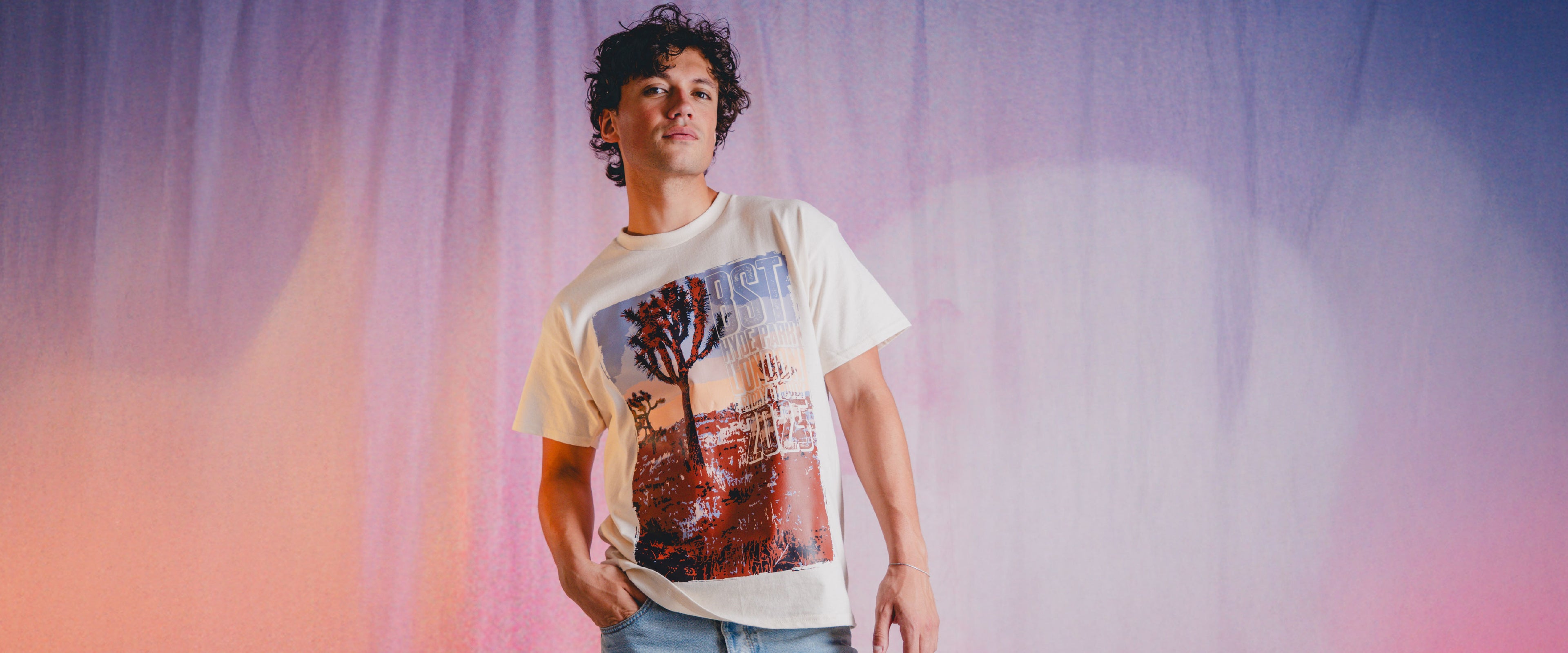A person with curly hair wearing a t-shirt with a colorful graphic print and light blue jeans stands against a gradient background of purple and pink hues.