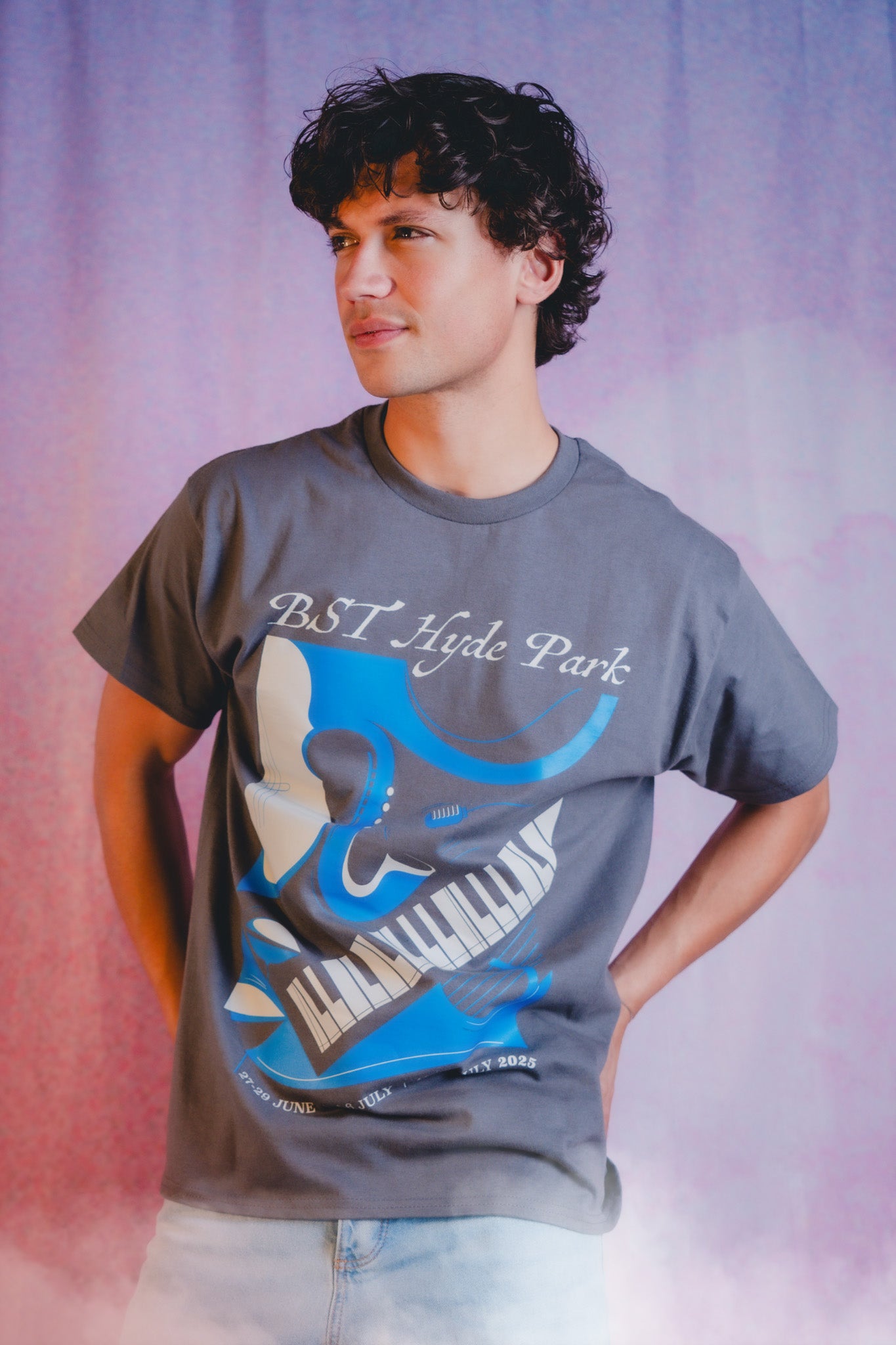 A person with curly dark hair wearing a gray T-shirt featuring a blue and white piano graphic and the text BST Hyde Park. They are standing with hands on hips against a pink and purple background.